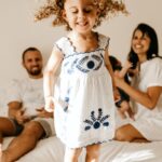 Adorable little girl with curly hair in light dress jumping on bed with closed eyes near happy young parents and newborn baby