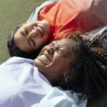 High angle of cheerful multiracial girlfriends lying on green lawn and toothy smiling while having fun together